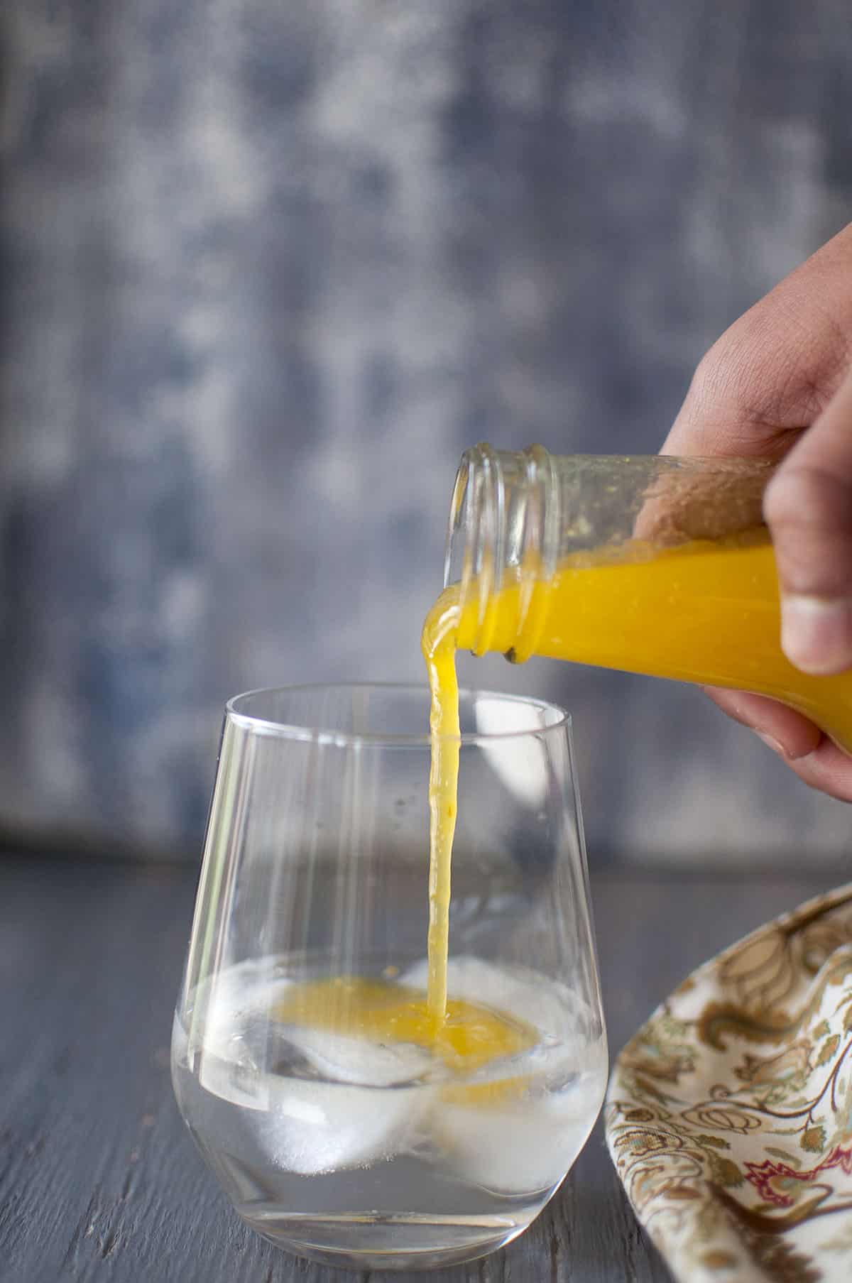 Hand holding a glass bottle with mango concentrate being poured into a glass with water and ice cubes