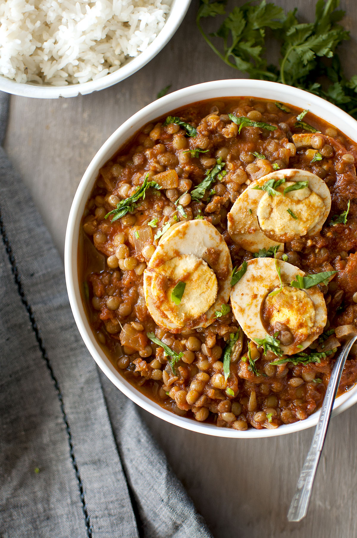 White bowl with lentils curry topped with egg halves and chopped cilantro served with a bowl of white rice