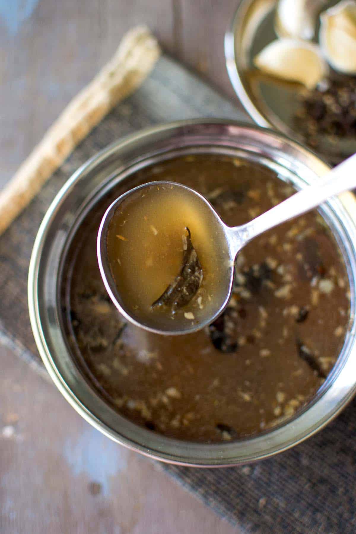 Hand holding a spoon of miriyala charu over a steel bowl.