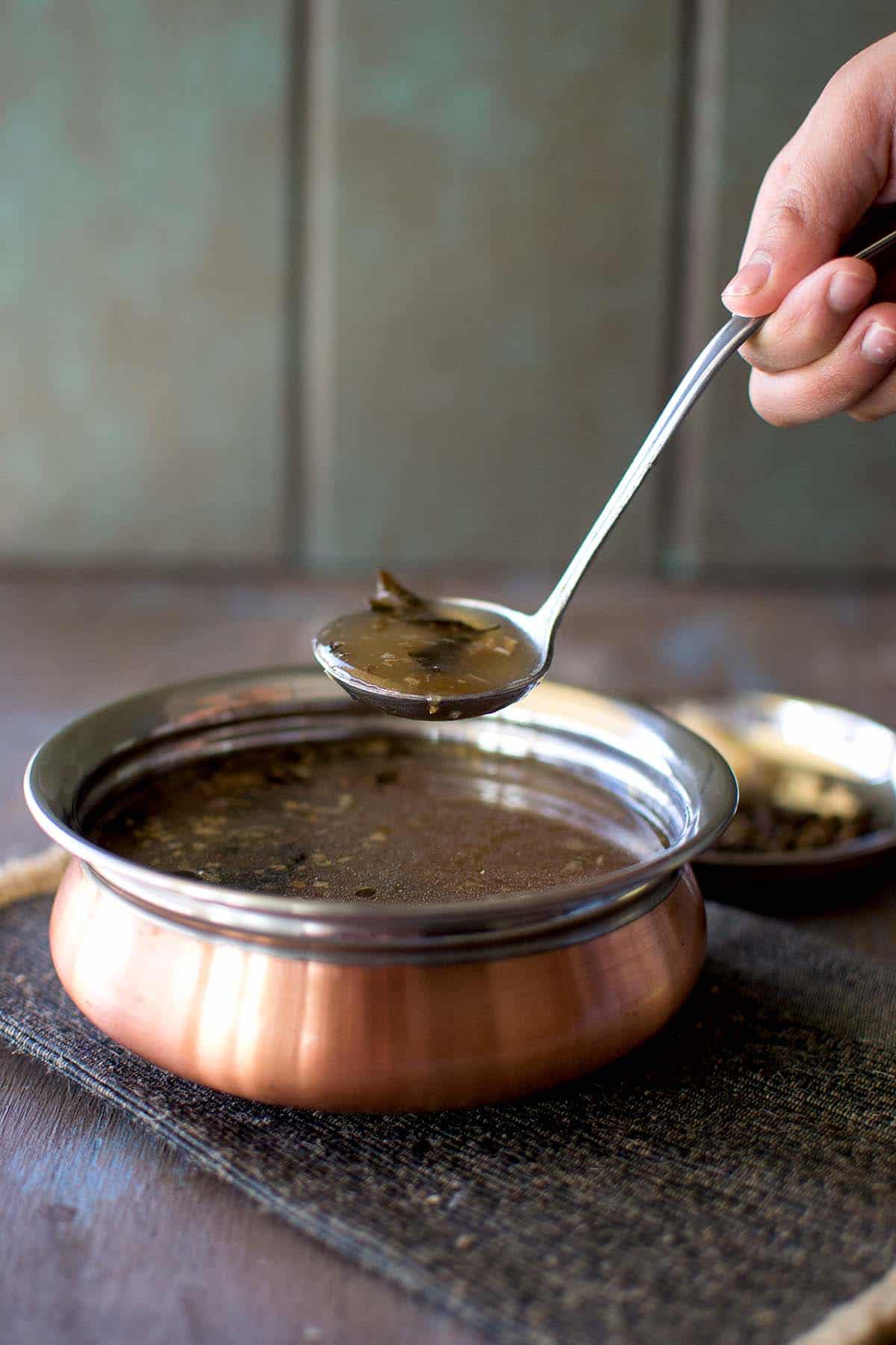 Hand holding a spoon over a bowl of pepper rasam.