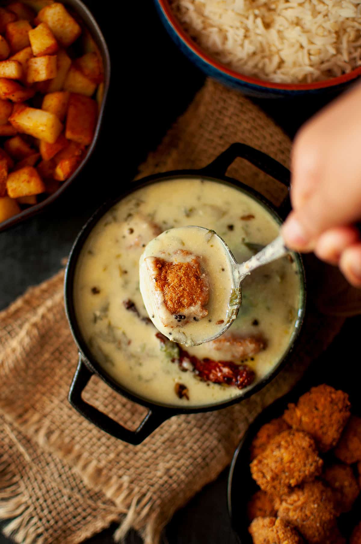 Hand holding a spoon with vada majjiga pulusu from a black pan.