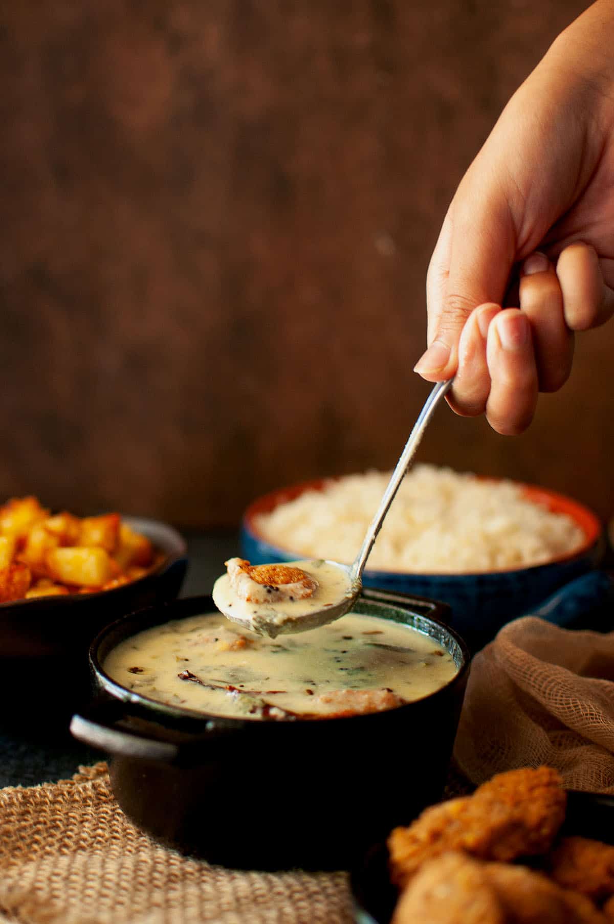 Hand holding a spoon with vada majjiga pulusu from a black pan.