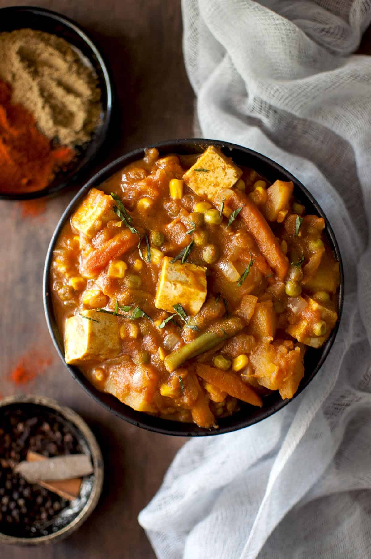 Top view of a black bowl with veg vindaloo and spices in the background