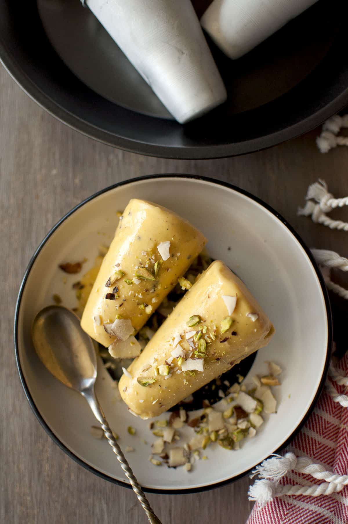 White plate with 2 mango kulfi pops topped with sliced almonds and pistachios