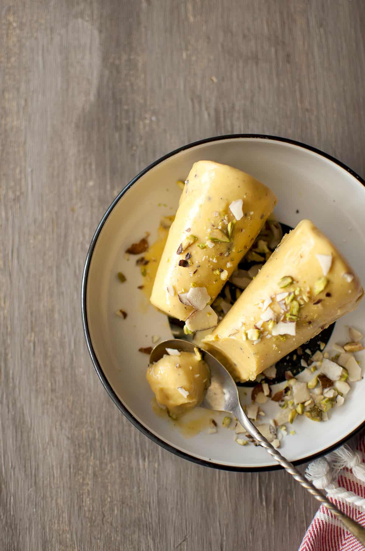 Black plate with 2 frozen popsicles topped with nuts and a spoon with a small scoop