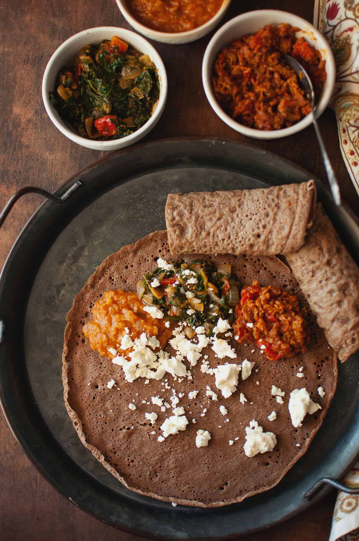 Top view of a tray with injera topped with side dishes and crumbled feta cheese.