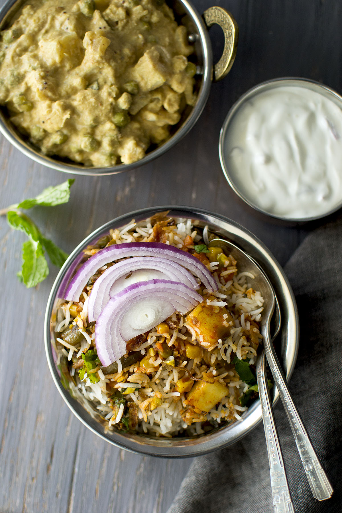 Steel bowl with rice topped with sliced onion and yogurt, curry in the background