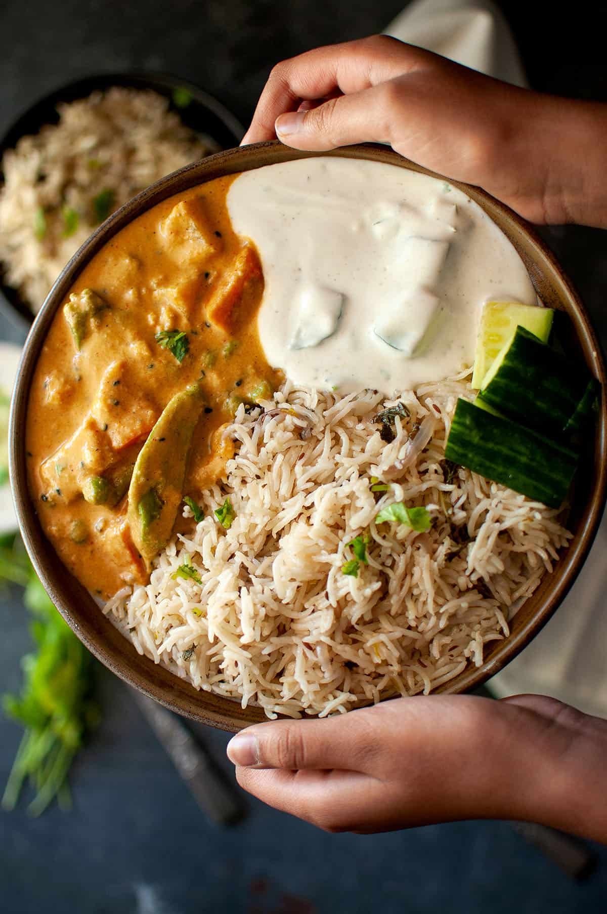 Hands holding a plate with salan, bagara rice, raita and cucumber.