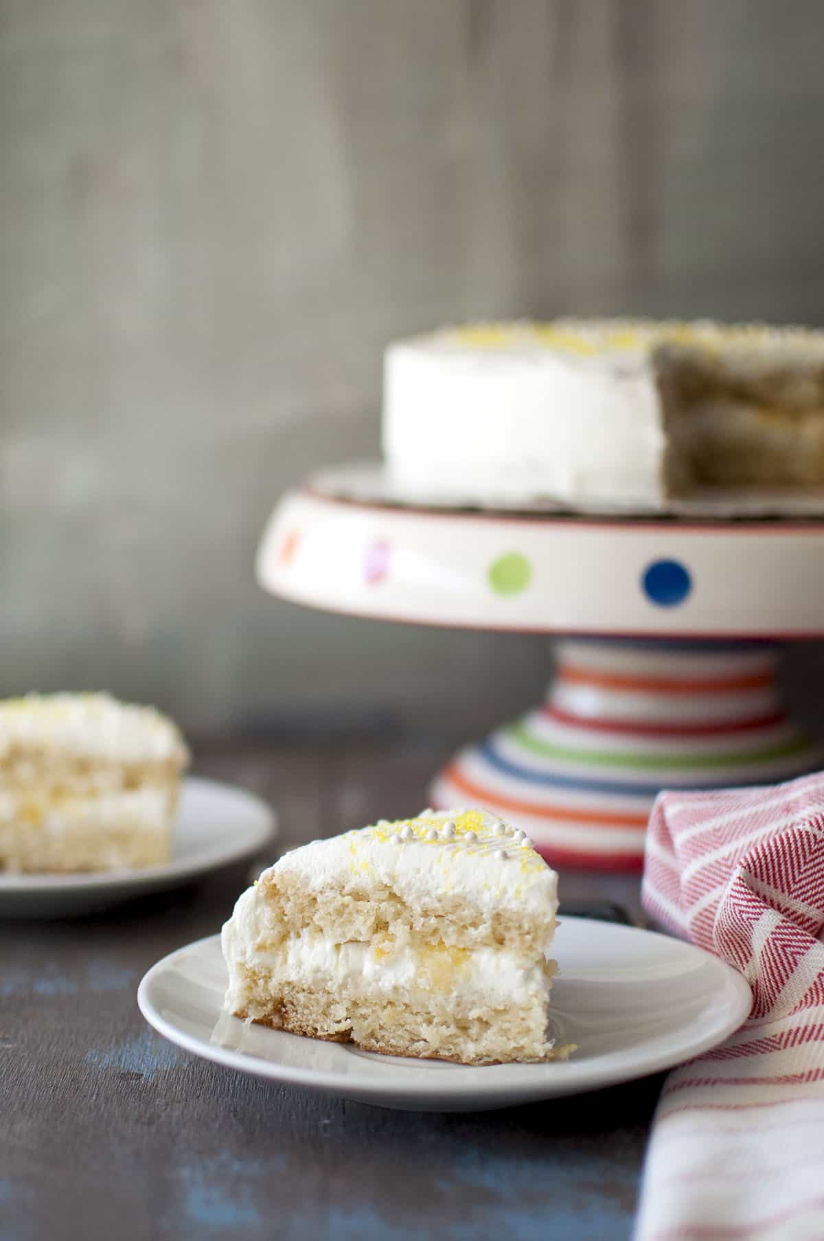 White plate with a slice of moist pineapple cake with a cake stand in the background