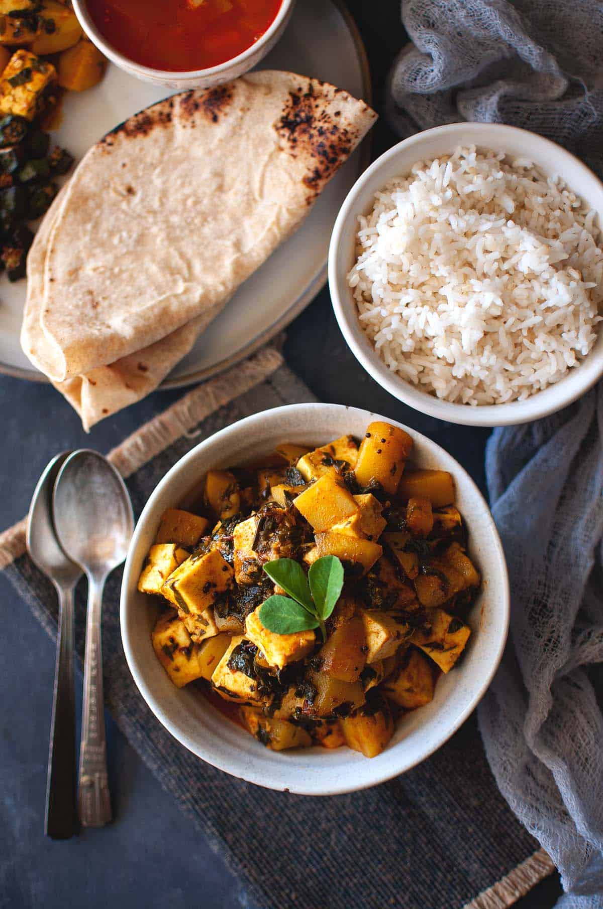 White bowl with aloo methi paneer curry with rice and roti in the background.