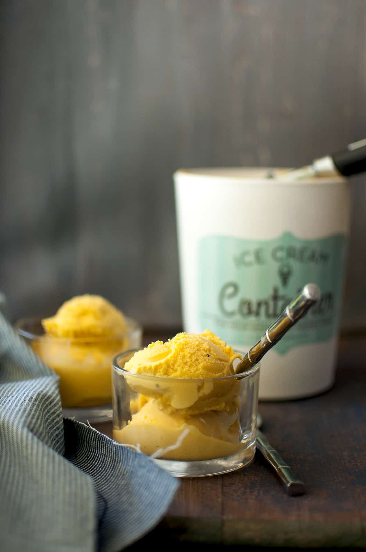 Glass serving bowl with 2 small scoops of mango gelato with a ice cream container in the background