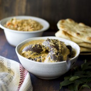 White bowl with bagara baingan with rice and naan in the background.