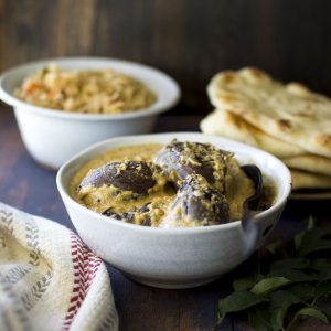 White bowl with bagara baingan with rice and naan in the background.