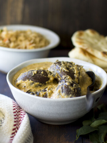 White bowl with bagara baingan with rice and naan in the background.