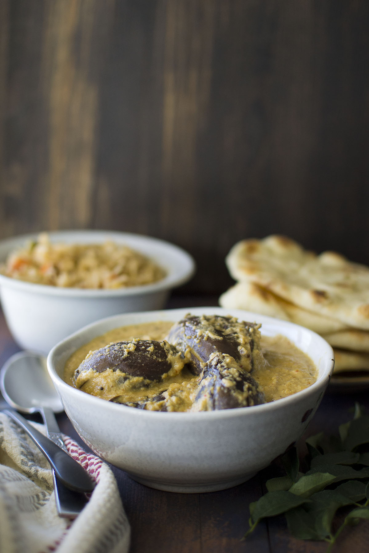 White bowl with bagara baingan with rice and naan in the background