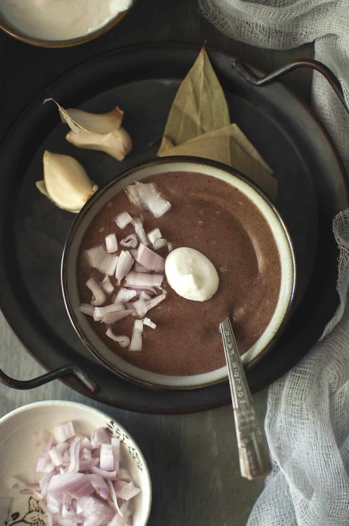 Black tray with a grey bowl of ragi porridge topped with chopped onion and yogurt