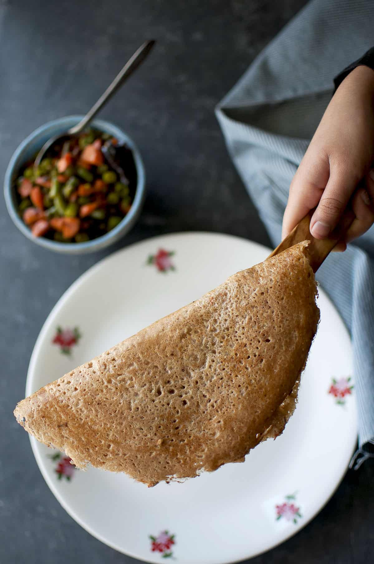 Hand holding a wooden spatula with wheat dosa