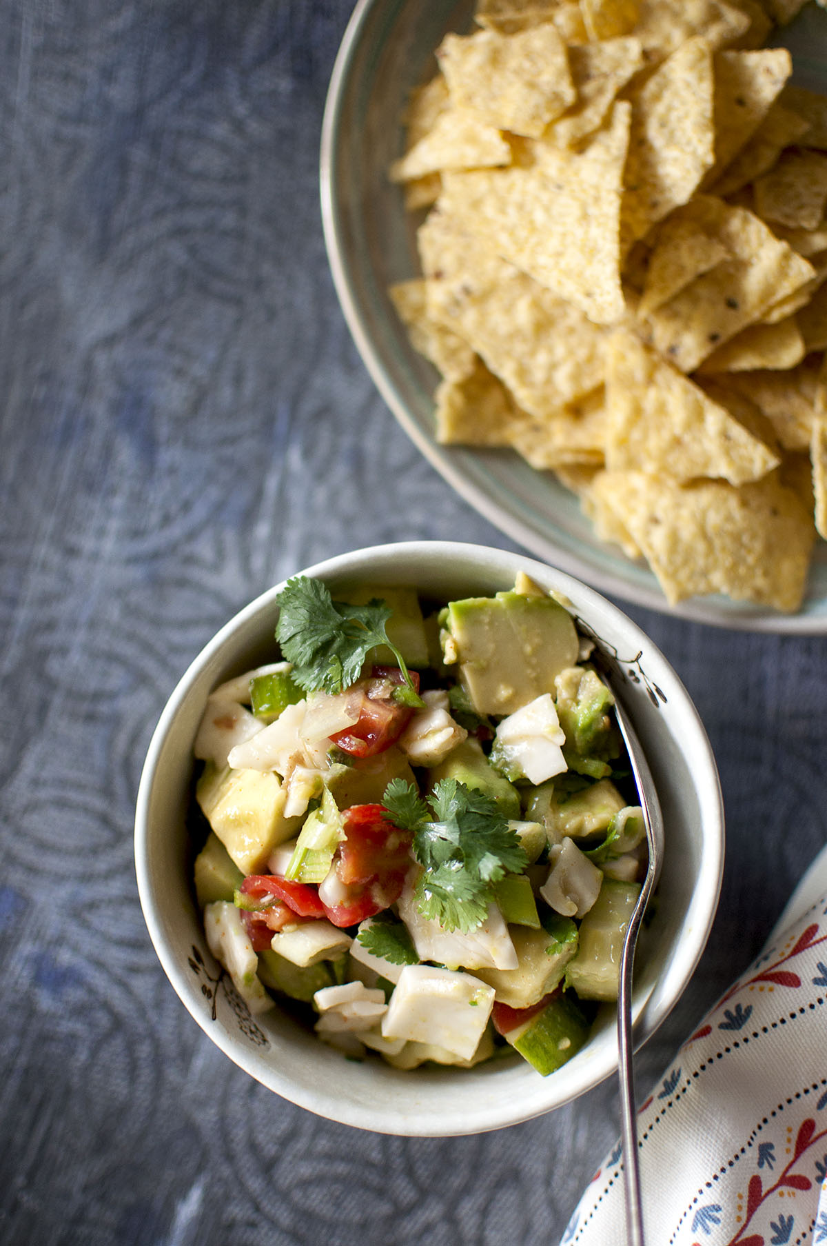 White bowl with chopped cucumber, avocado, tomatoes, coconut, cilantro and blue plate with tortilla chips