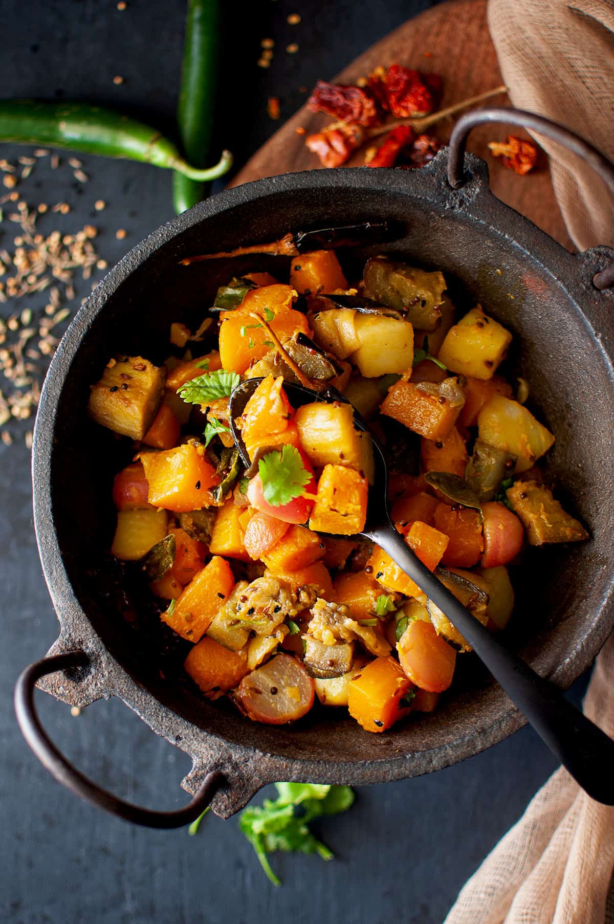 Cast iron wok with mix vegetable curry with a black spoon.