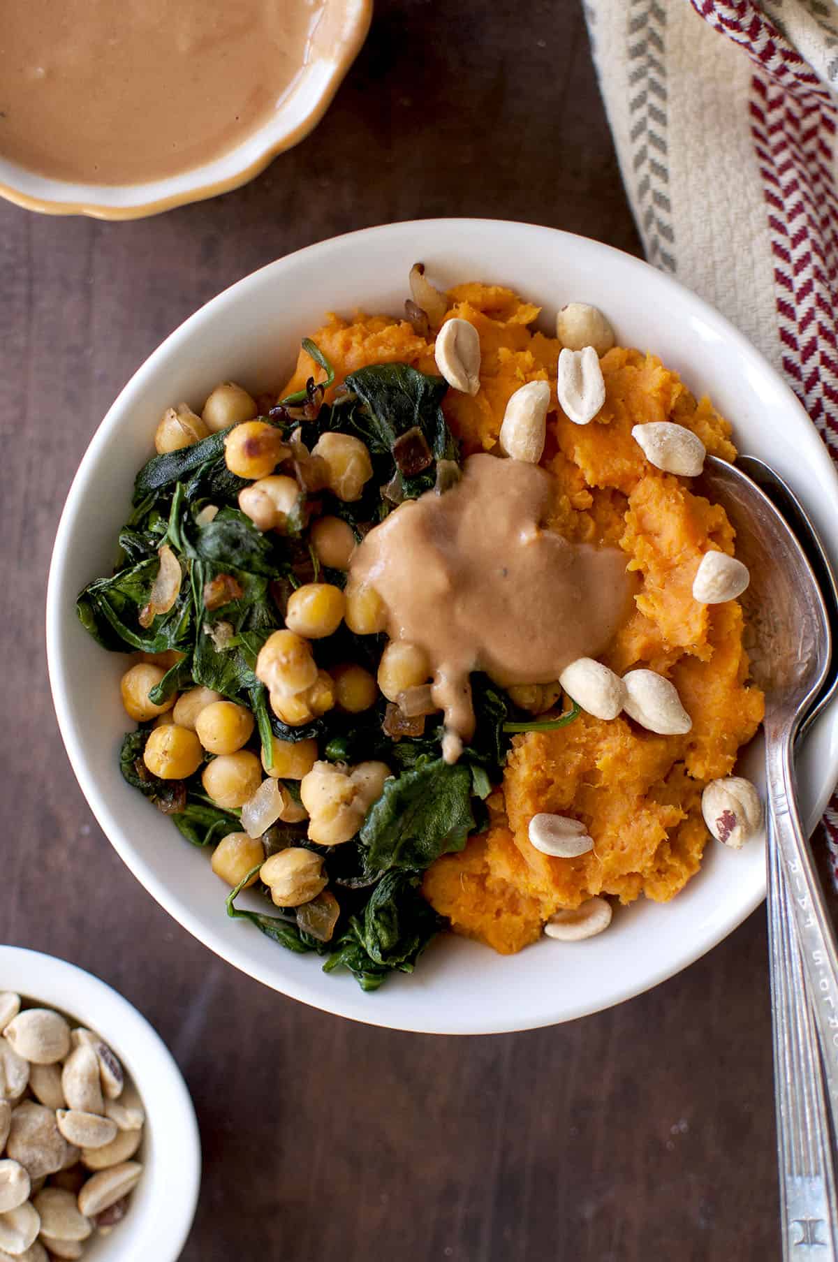 White bowl with mashed sweet potato, sautéed green, chickpeas and g-nut sauce