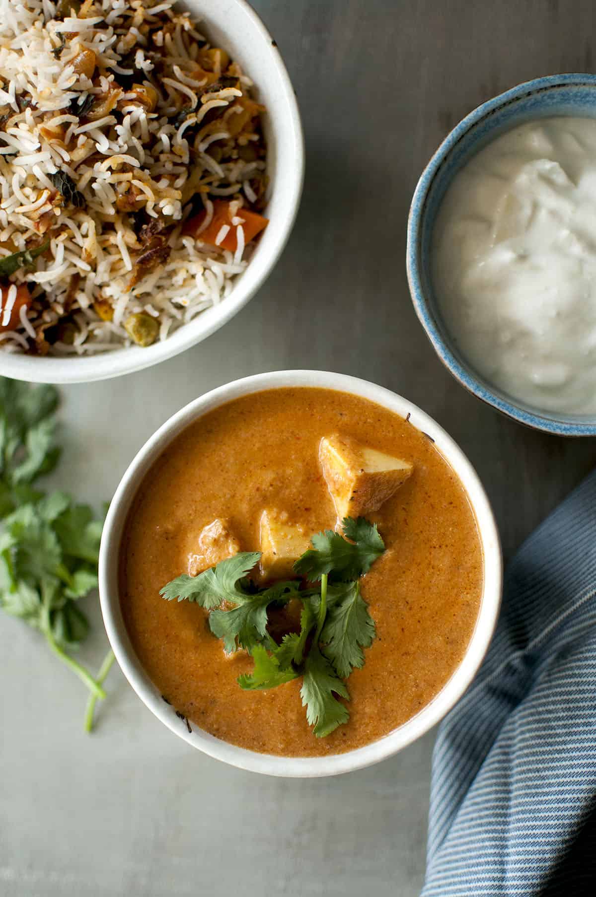 White bowl with paneer tikka masala with a rice pilaf and yogurt in the background