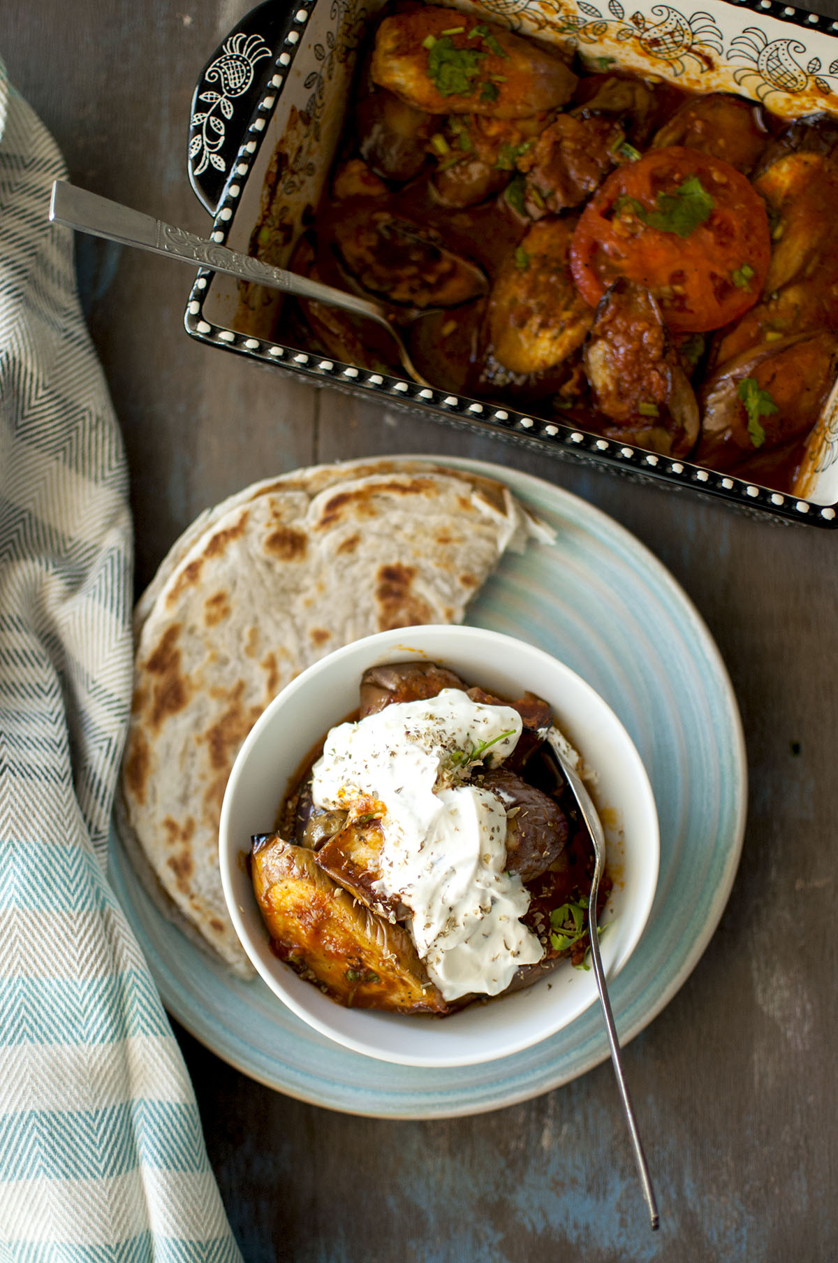 White bowl with borani banjan topped with garlic yogurt and served with flatbread on the side.