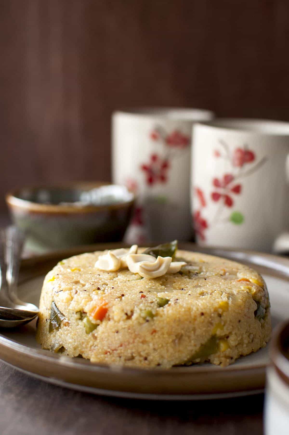 brown plate with jonna rava upma topped with cashews and curry leaves and 2 coffee mugs in the background