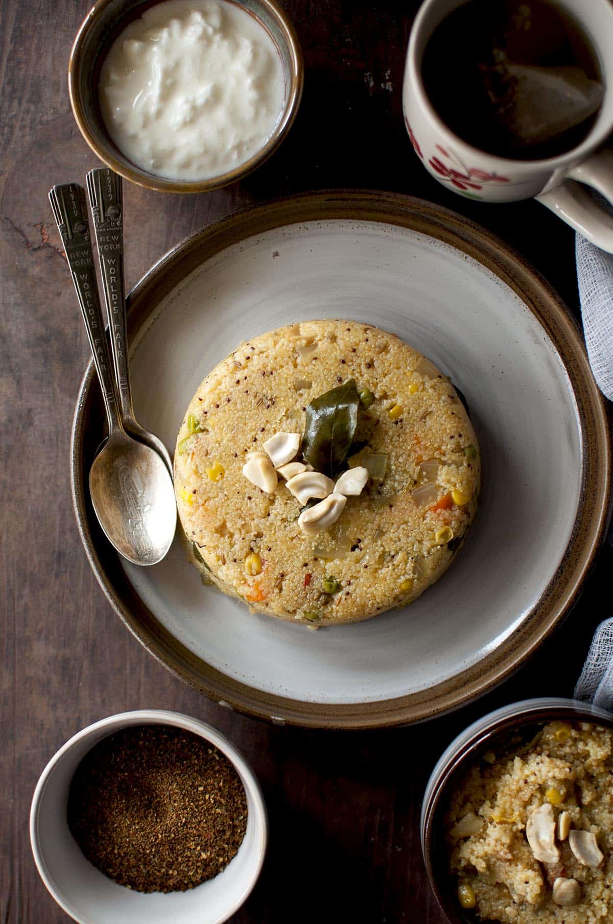 Grey plate with jonna rava upma and 2 spoons and a bowl of yogurt in the background