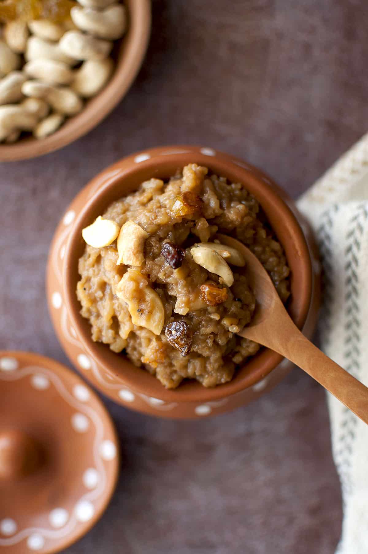 Terracotta pot with sweet pongal topped with raisins and cashews and a wooden spoon