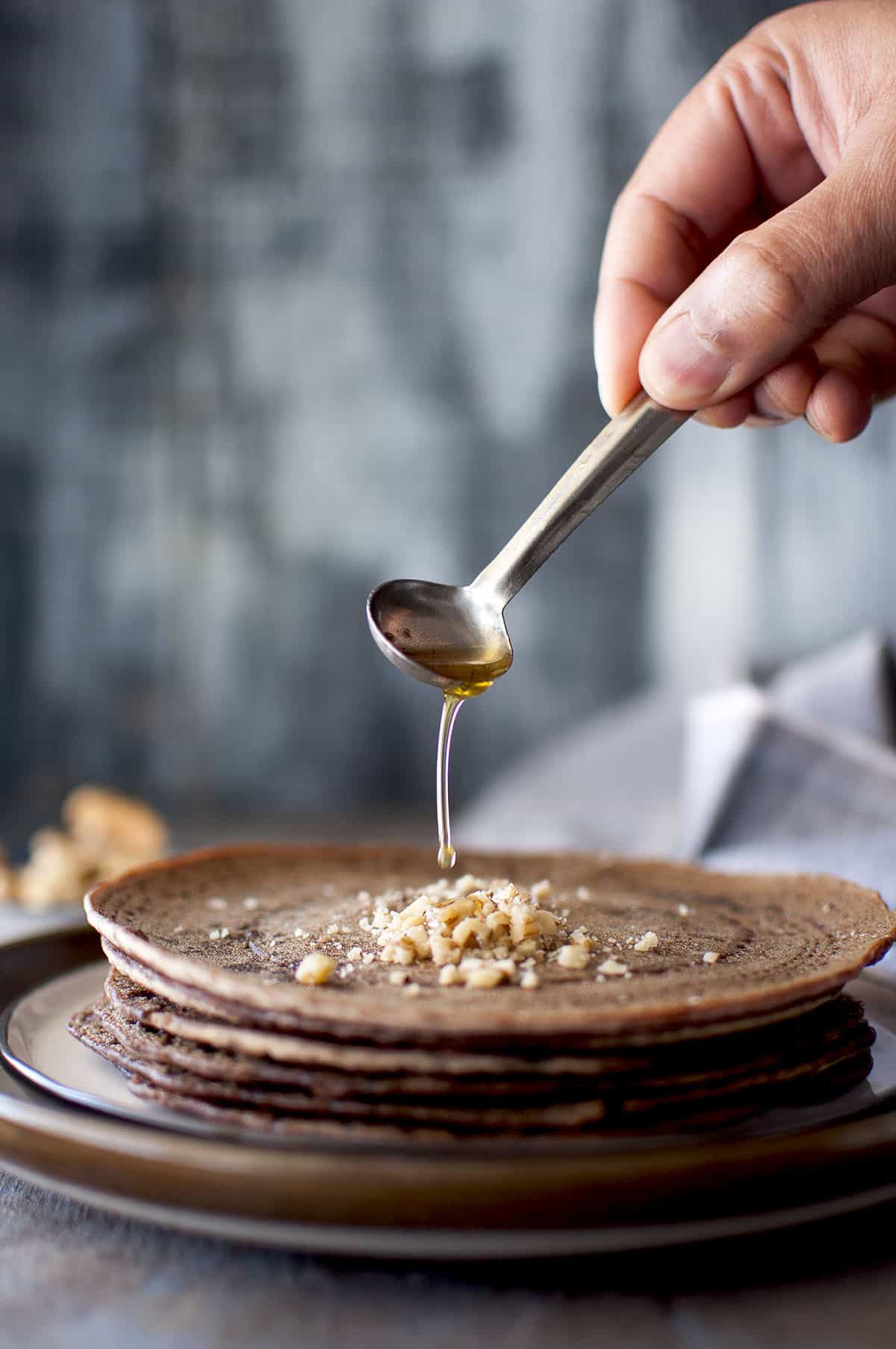 Hand holding a steel spoon and drizzling melted ghee onto a stack of ragi dosa