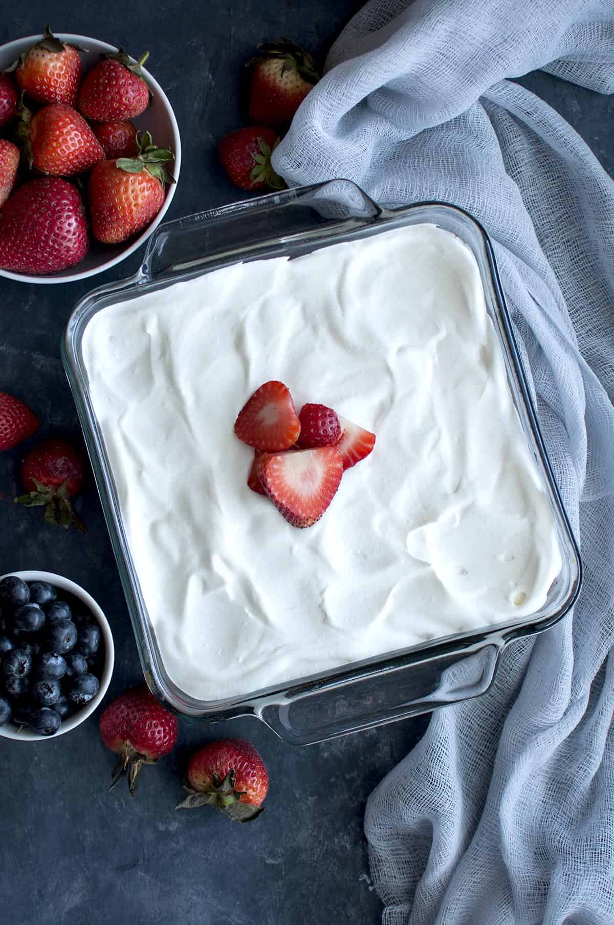 Glass square baking pan with whipped cream and sliced strawberry topped cake