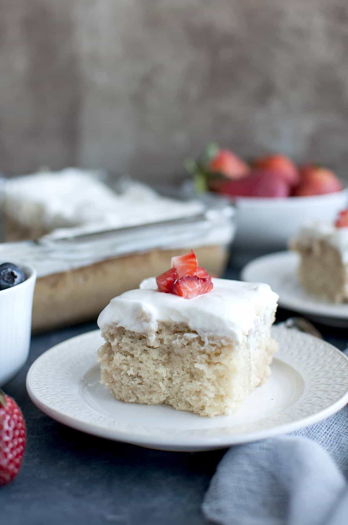 White plate with a slice of vegan tres leche cake topped with whipped cream and chopped berries