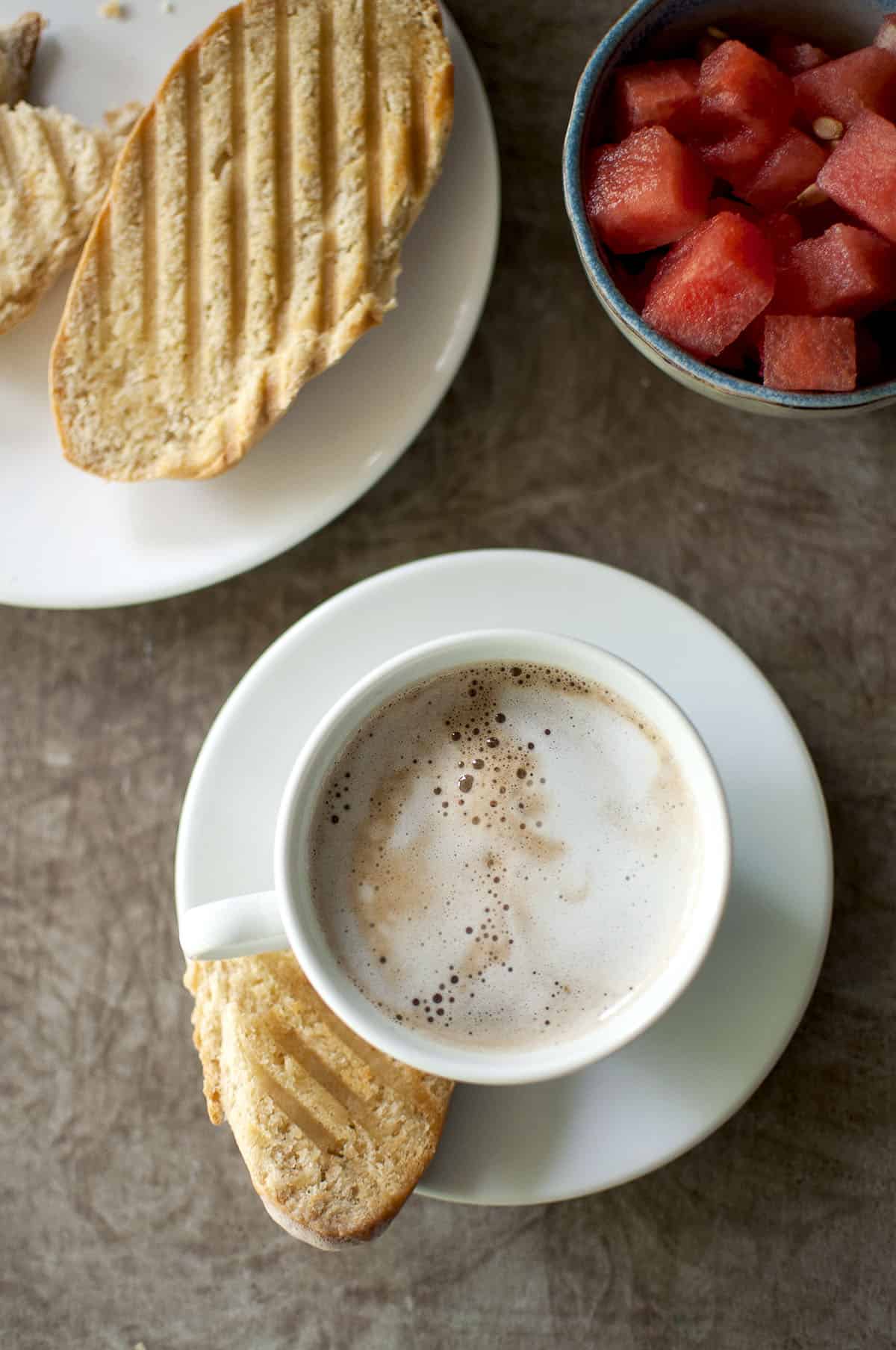 White cup with frothy cafe con leche and a piece of toast with a bowl of watermelon