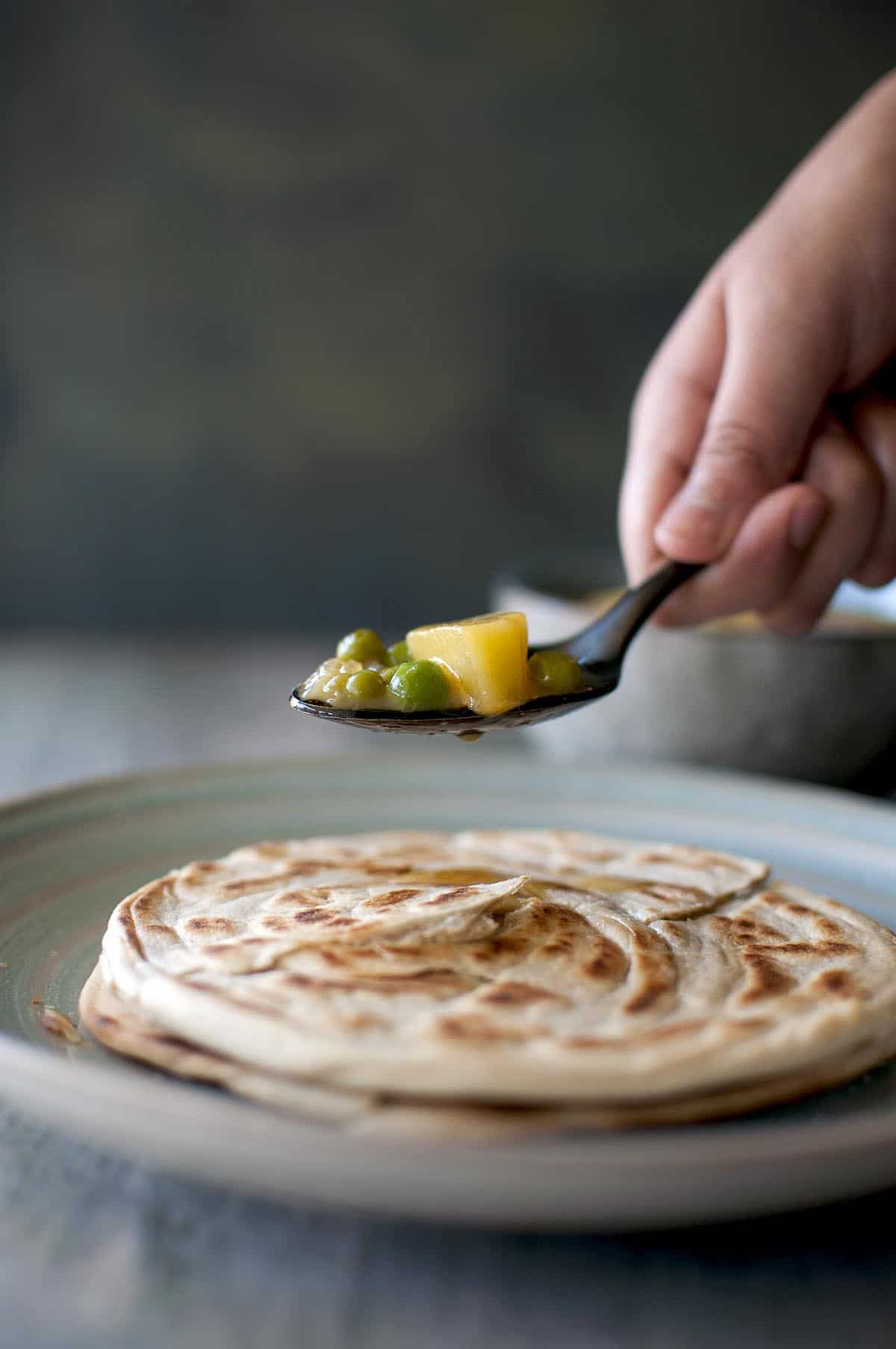 Hand holding a black spoon with Malaysian curry sauce over roti canai