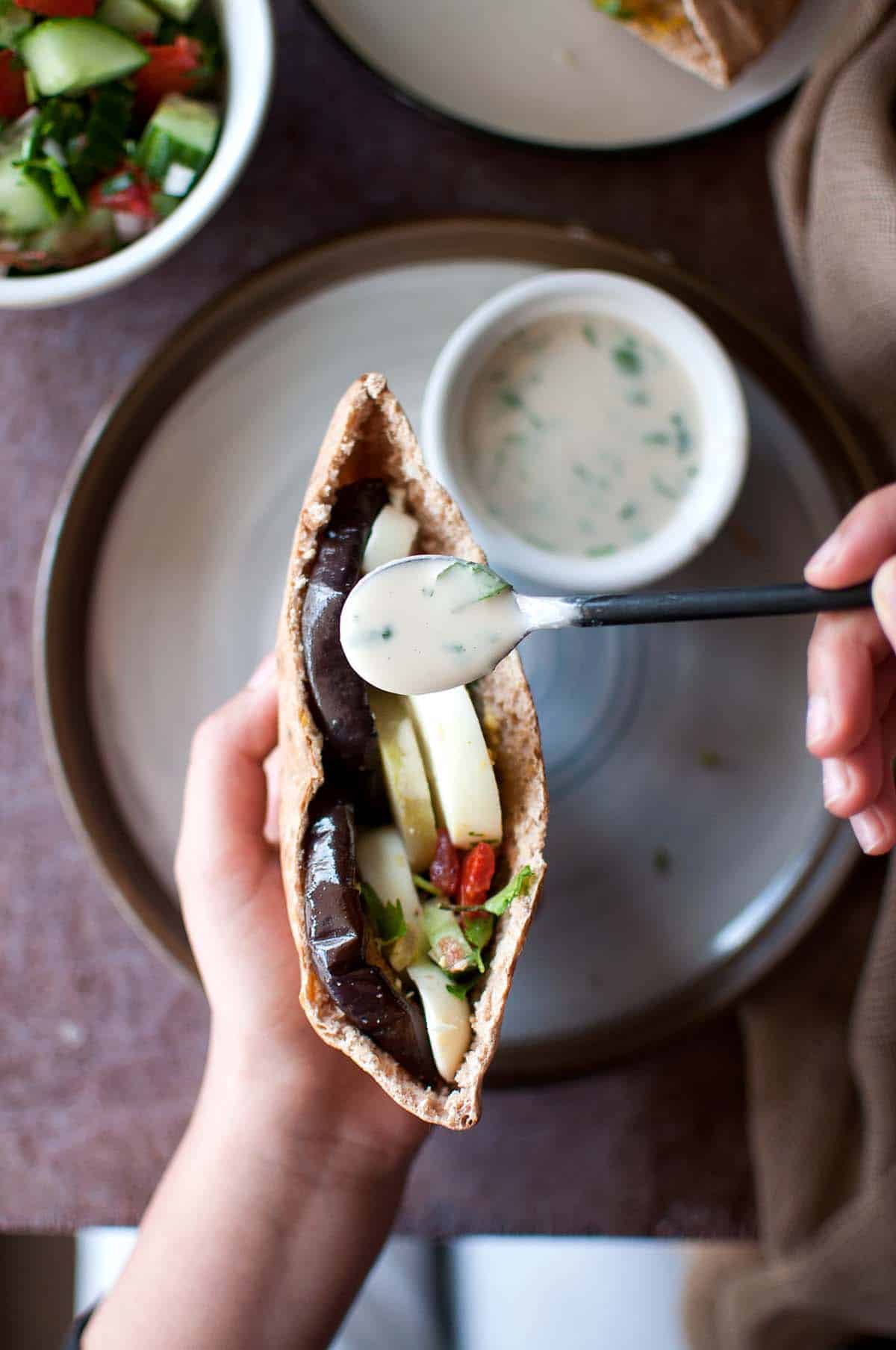 Hand pouring tahini sauce on a pita sandwich.