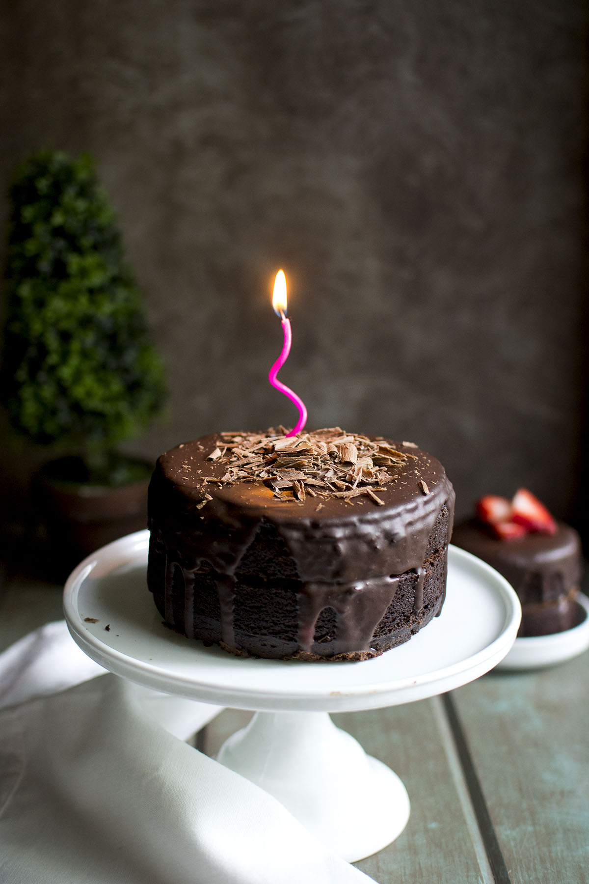 White cake stand with vegan chocolate cake with a lit candle on top