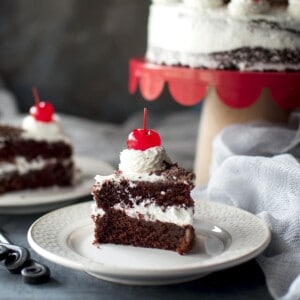 White plate with a slice of 2 layered chocolate cake and topped with cherry