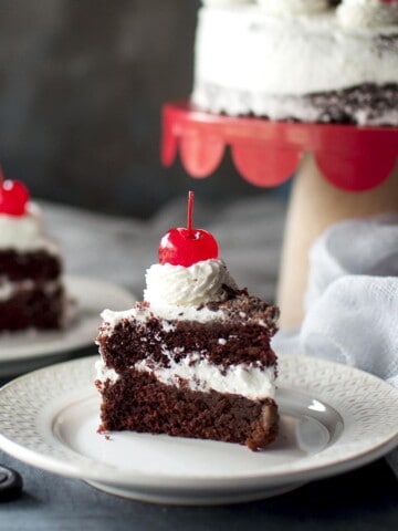 White plate with a slice of 2 layered chocolate cake and topped with cherry