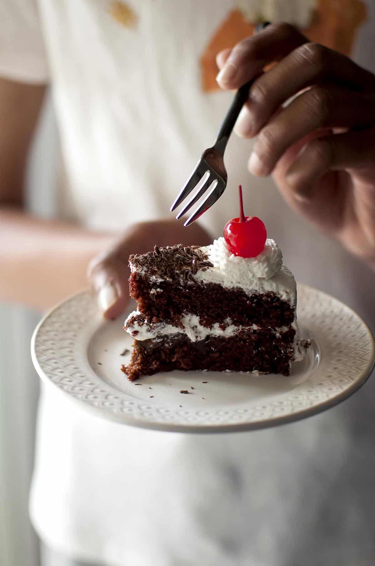hand holding white plate with a slice of black forest cake and a fork in another hand