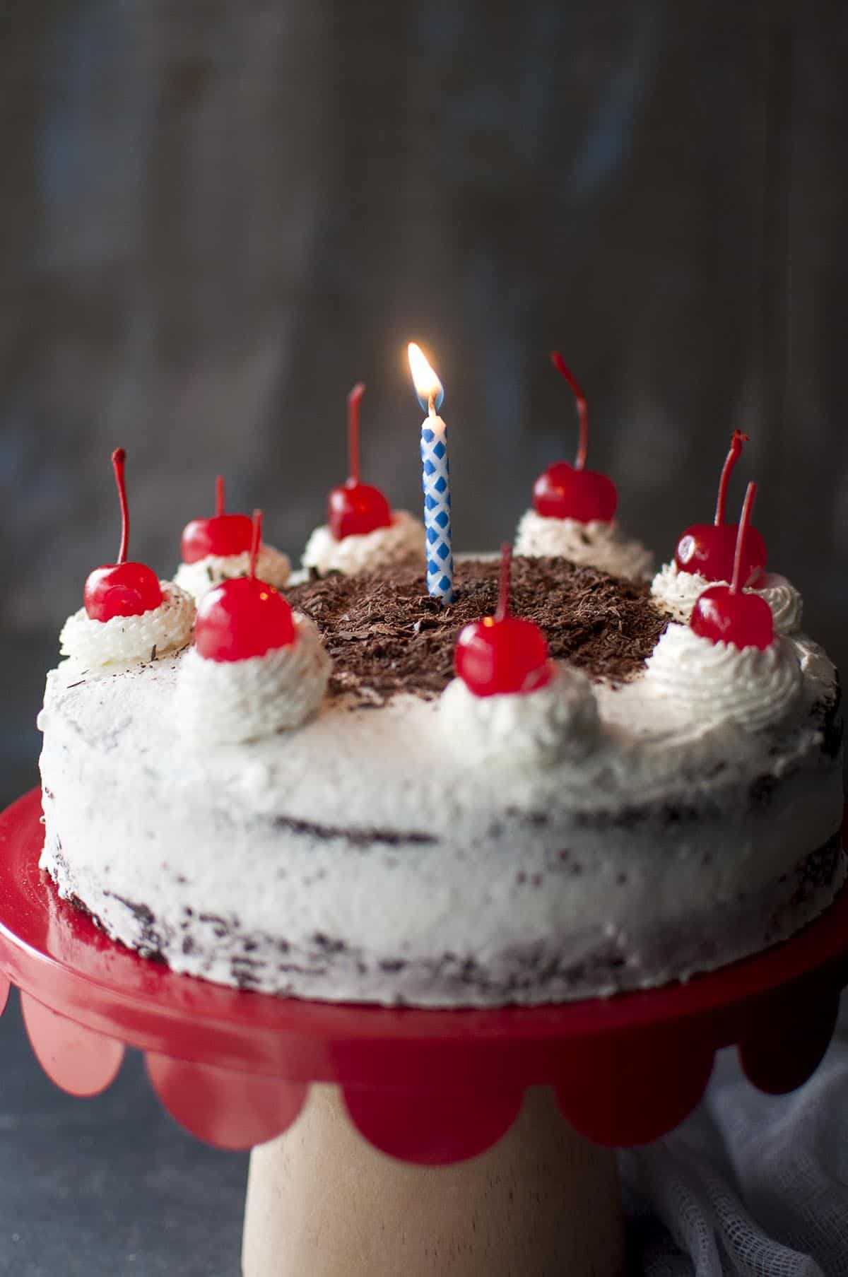 Red scallop cake stand with 2 layer eggless chocolate cake topped with a lit candle