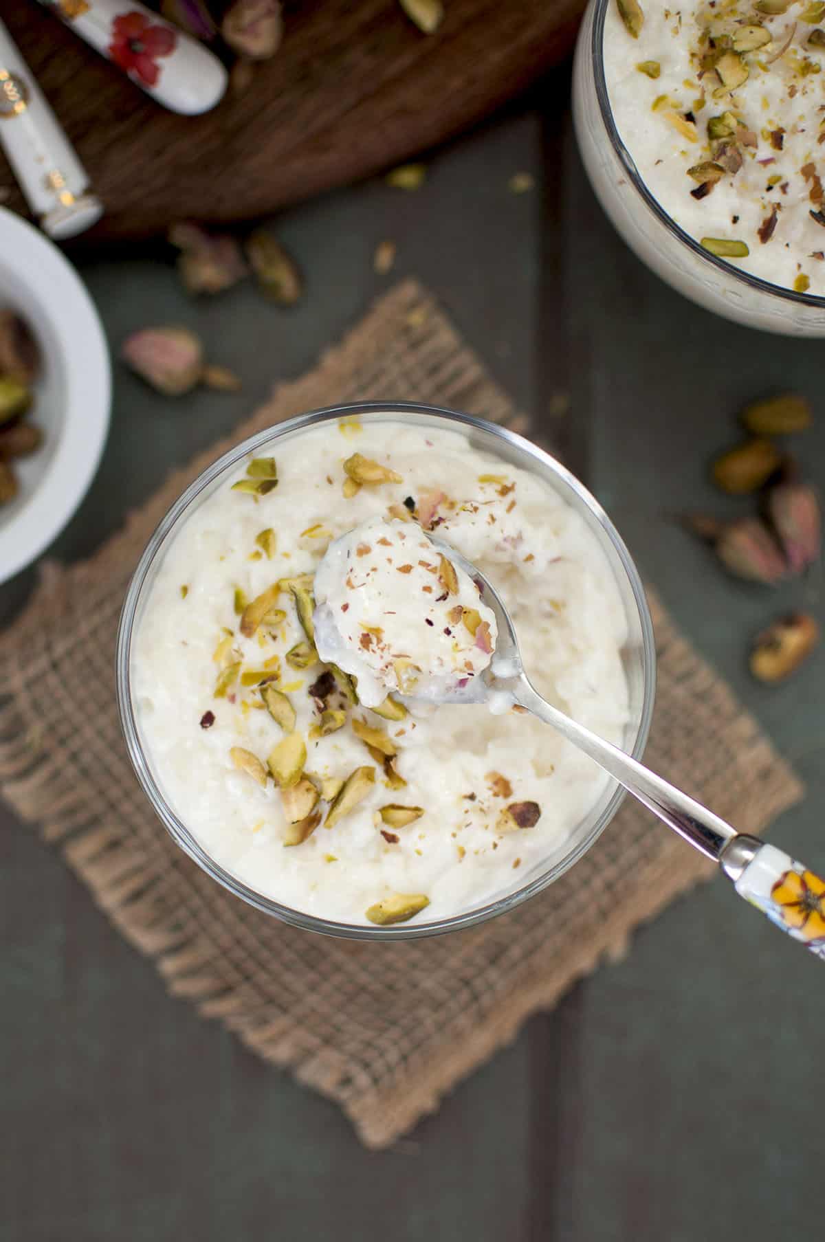 Glass serving cup with rice pudding topped with chopped pistachios and dried rose buds and a spoon on top