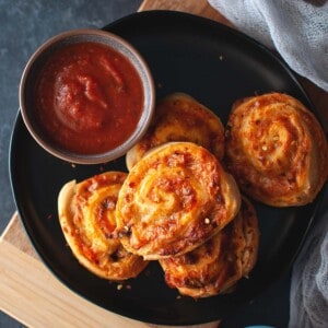 Black plate with a stack of pizza pinwheels and a bowl of marinara sauce.