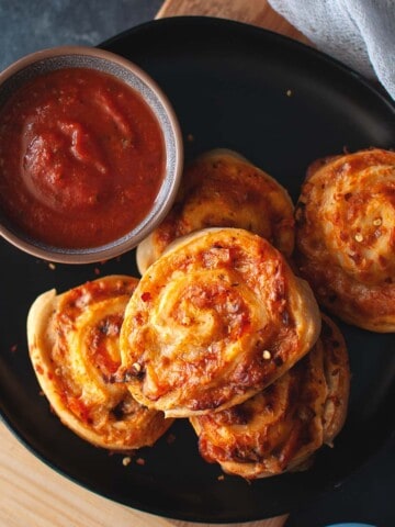 Black plate with a stack of pizza pinwheels and a bowl of marinara sauce.