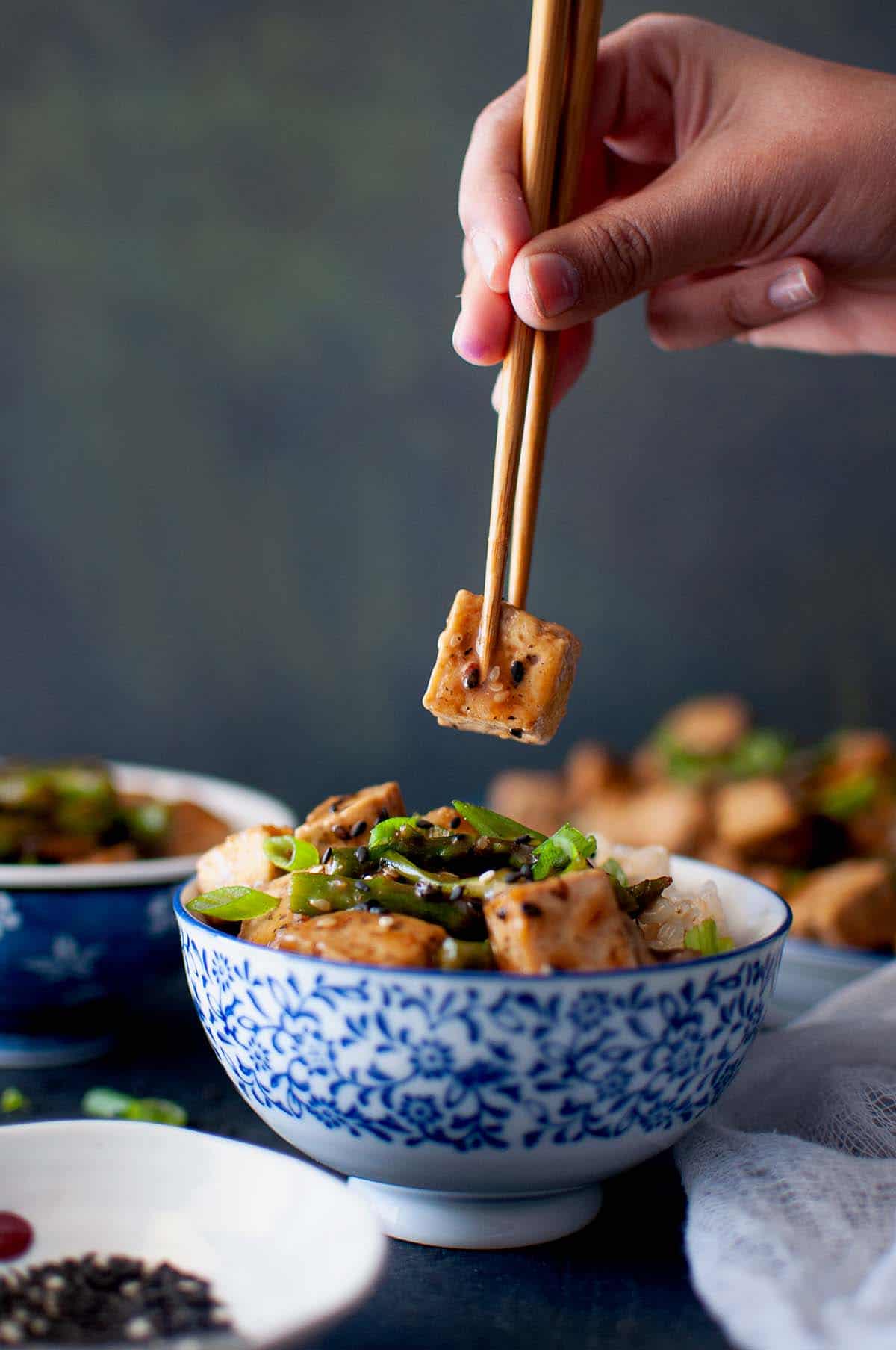 Hand holding a cube of tofu with chopsticks.