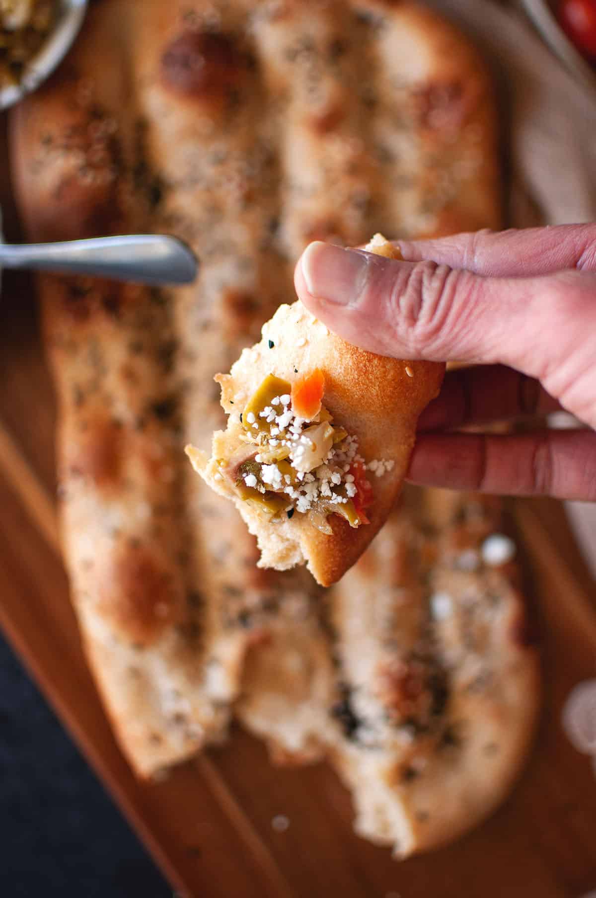 Hand holding a piece of barbari bread topped with cheese and olives.