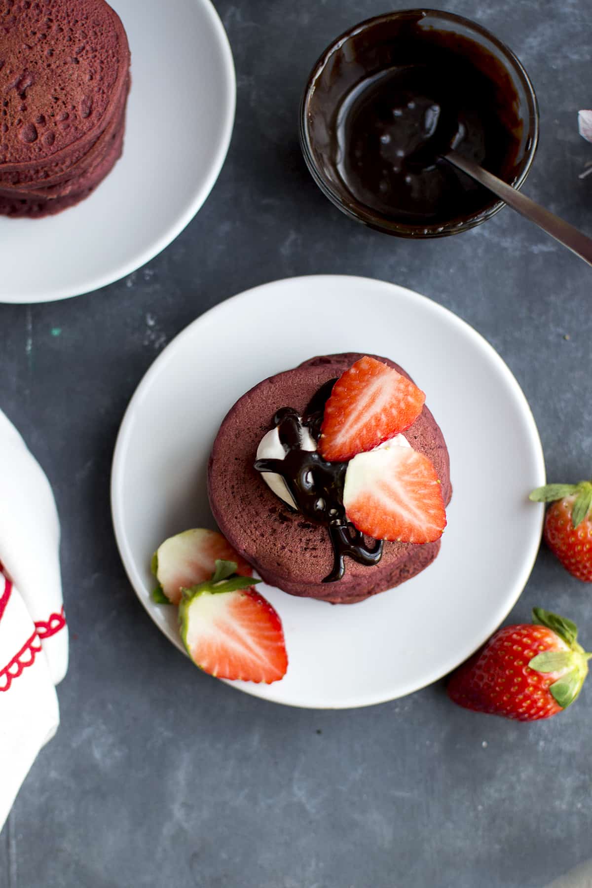 White plate with a stack of red velvet pancakes and a bowl of chocolate syrup in the background