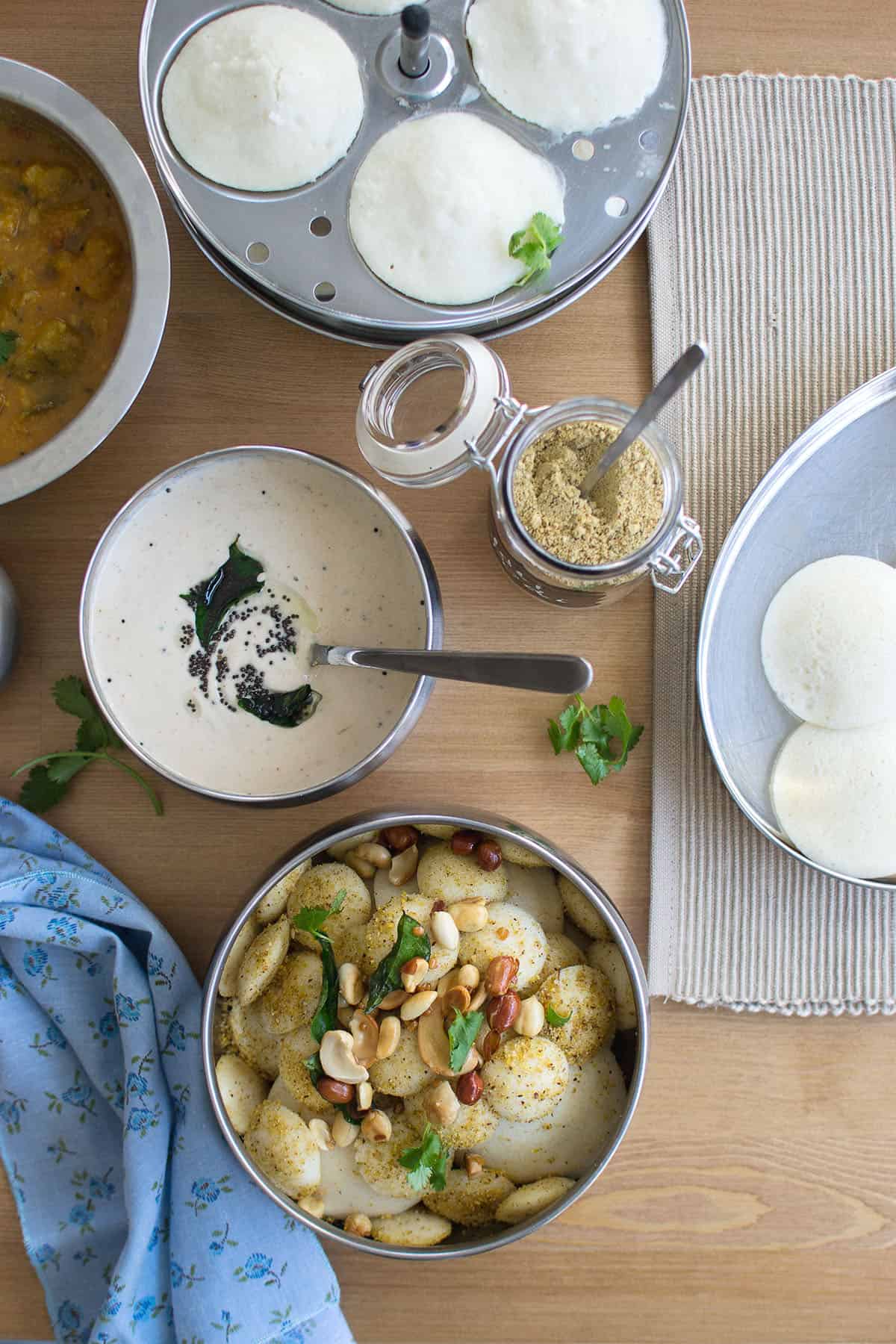 Top view of a dining table with steel bowl with podi Idli with chutney and sambar in the background.