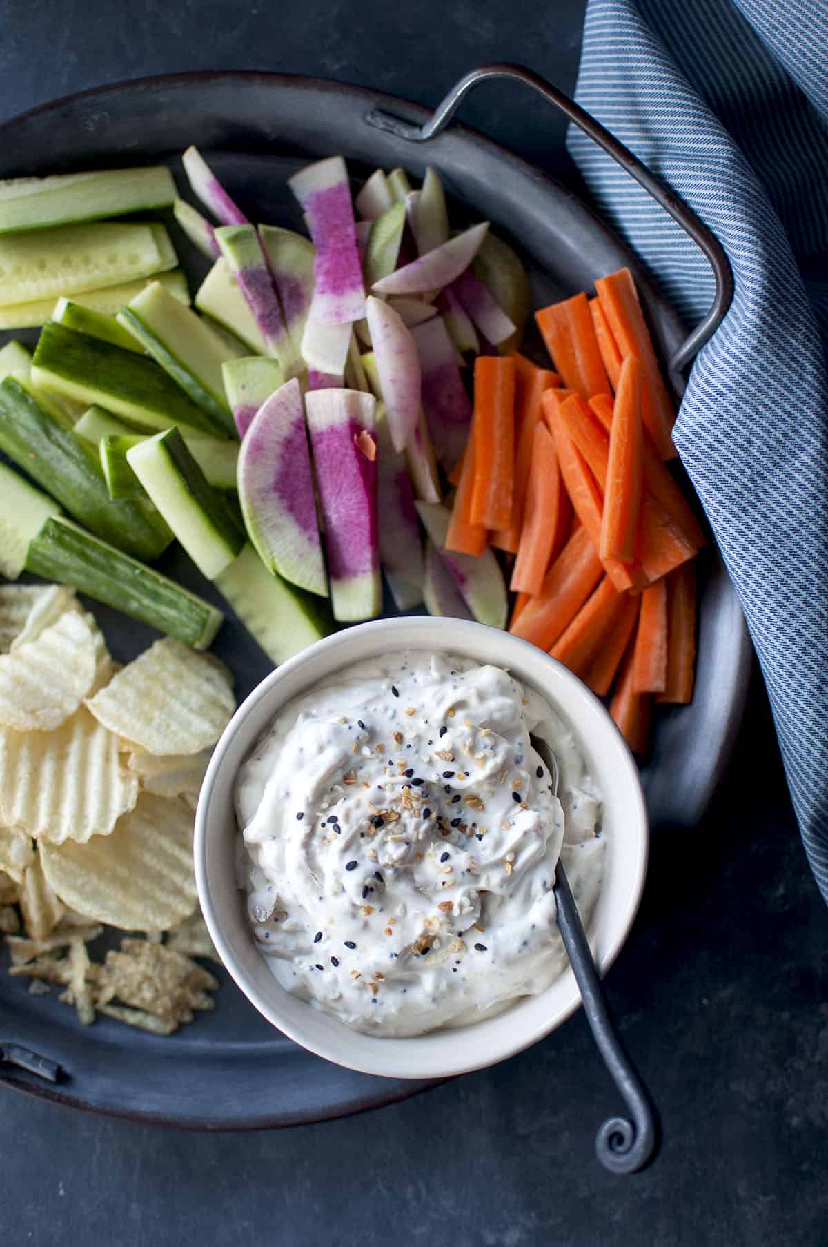 Tray with chopped veggies and a white bowl with bagel dip and a spoon