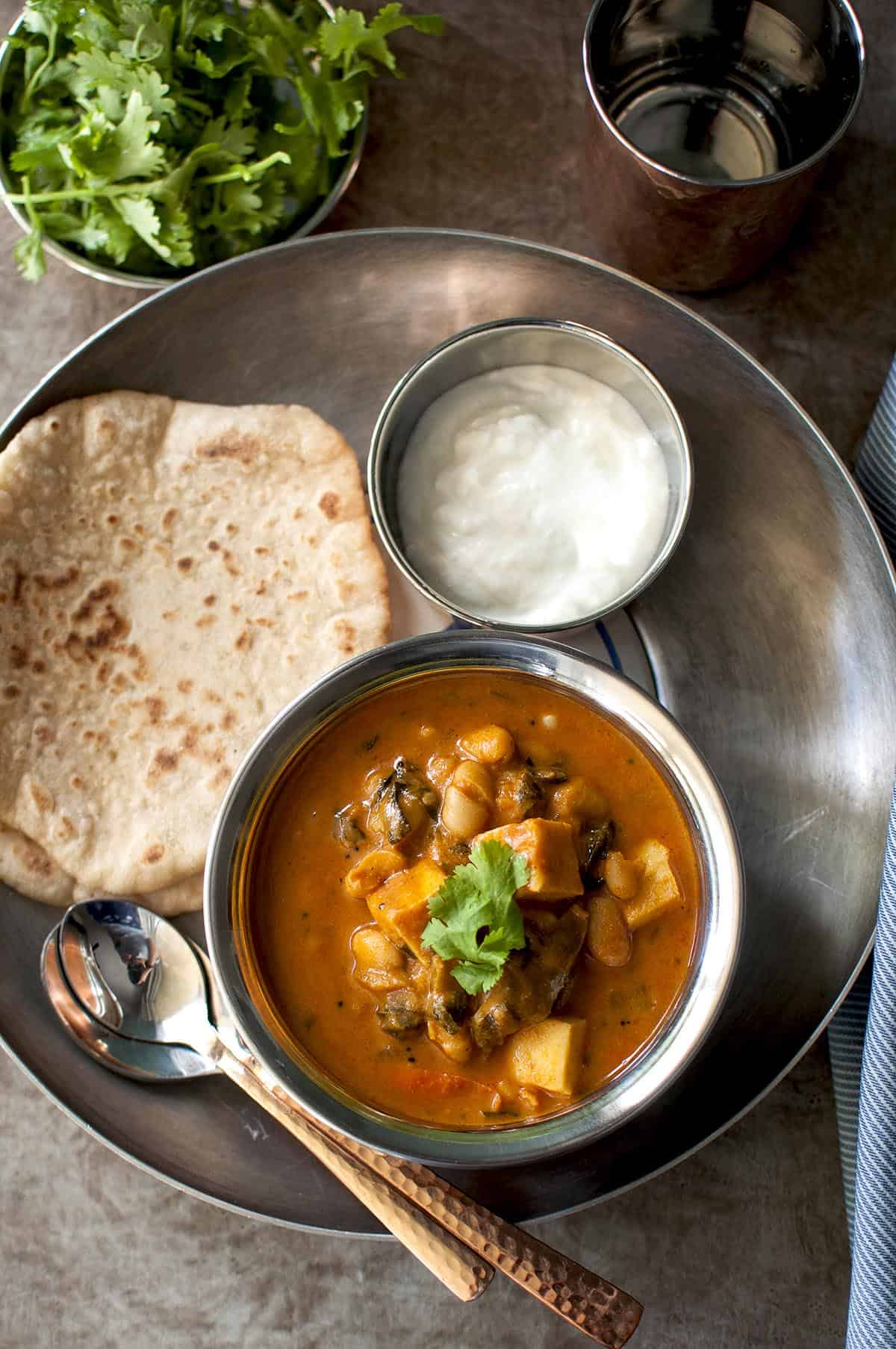 Tray with steel bowl of paneer curry, flatbread and a bowl of yogurt