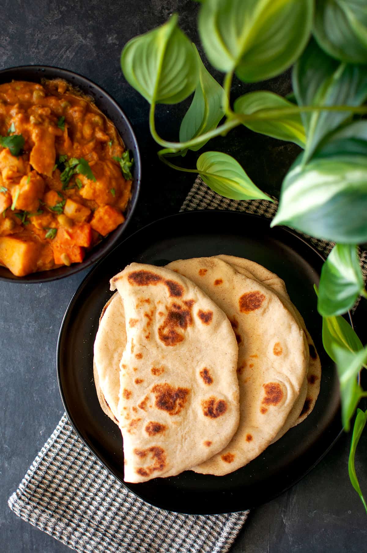 Black plate with a stack of Yogurt flatbread and a black bowl with curry in the back.